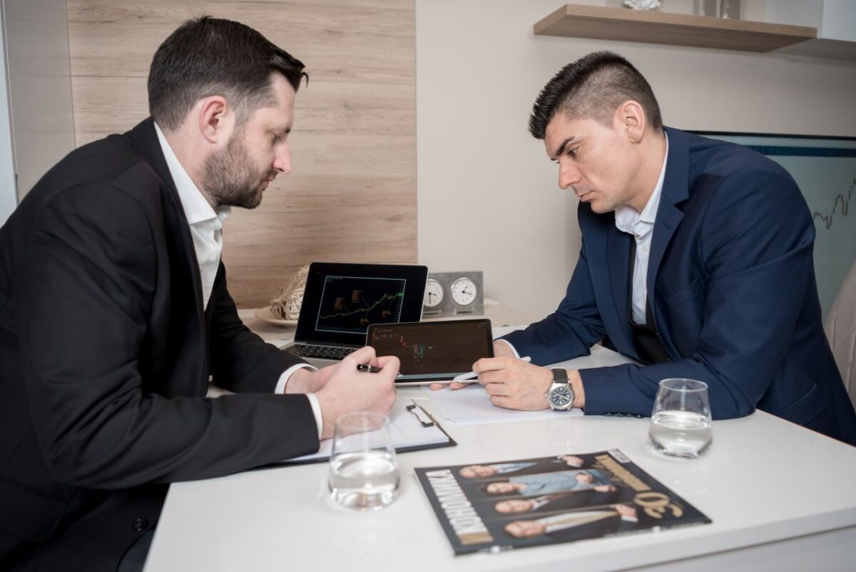Promotion Rue 66 Two businessmen in suits discussing cryptocurrency trends using digital devices in an office setting.